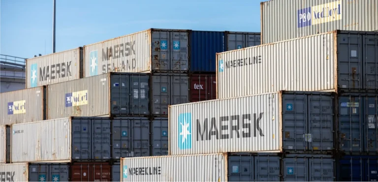 Stacked shipping containers in a yard, showing Maersk and Nedlloyd logos against a blue sky.