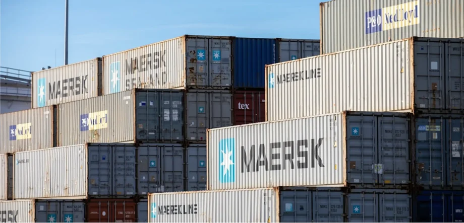 Stacked shipping containers in a yard, showing Maersk and Nedlloyd logos against a blue sky.